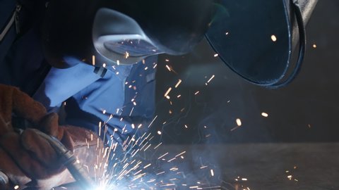 Close-up blacksmith welder in protective mask works with metal using a welding machine, bright sparks and flashes in slow motion