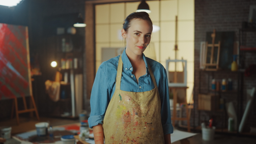 Portrait of Talented Young Female Artist Dirty with Paint, Wearing Apron, Crosses Arms while Holding Brushes, Looks at the Camera with a Smile. Authentic Creative Studio with Large Canvas and Tools  Royalty-Free Stock Footage #1036269968
