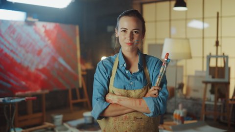 Portrait of Talented Young Female Artist Dirty with Paint, Wearing Apron, Crosses Arms while Holding Brushes, Looks at the Camera with a Smile. Authentic Creative Studio with Large Canvas and Tools 