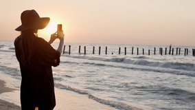 Girl in a black hat filming beautiful sunset on a seaside. Summer warm golden hour light, evening landscape and sea. Yellow sand and calm waves. Woman making selfie photos and videos on telephone 