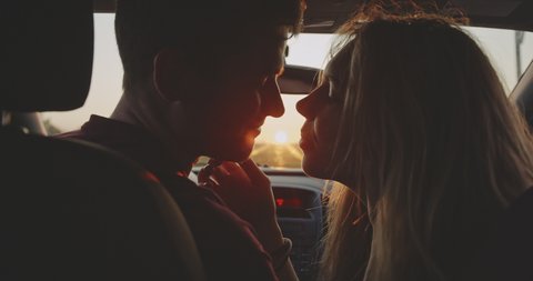 Romantic young couple kiss on the front sit of their car during the road trip. Beautiful young woman kissing her boyfriend at sunset golden light at first date. Concept of love, relations and travel.