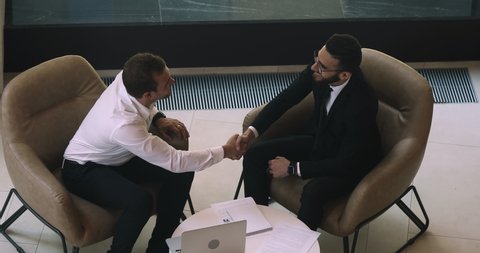 Happy diverse male business partners talk handshake sit on chair at office meeting, two businessmen shake hands making business agreement establish partnership investment deal concept, above top view