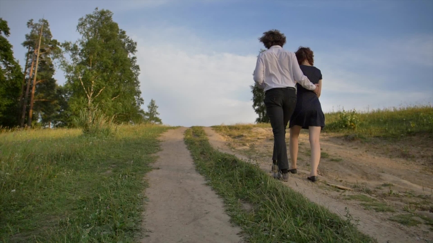Two young men a guy and a girl hugging climb the mountain, along the path in the park. Blue sky and green trees on a warm summer day. Pleasant evening walk Concept.