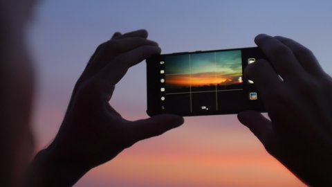 Male hands holding mobile phone with image of sunset or sunrise on the screen taking photo of picturesque scenery