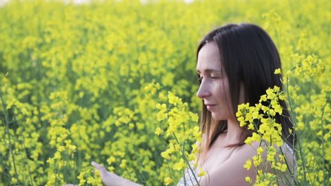 A beautiful carefree girl with healthy hair and freckles on her face against a yellow rapeseed field. Attractive brunette in white dress enjoys yellow rape flowers, natural beauty.