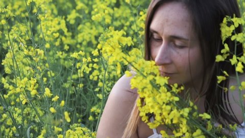 A beautiful carefree girl with healthy hair and freckles on her face against a yellow rapeseed field. Attractive brunette in white dress enjoys and sniffs yellow rape flowers, natural beauty