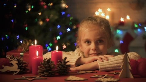 Little girl looking at Christmas decorations on table, lights twinkling on tree