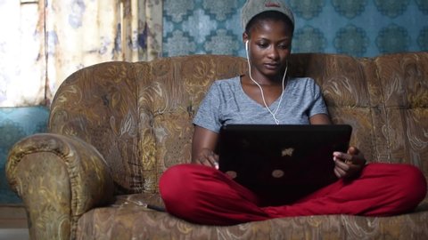 Auchi, Edo State/ Nigeria - August 29 2019: beautiful african girl sitting on a couch using her laptop, picks up her phone, checks it, and gets excited and happy