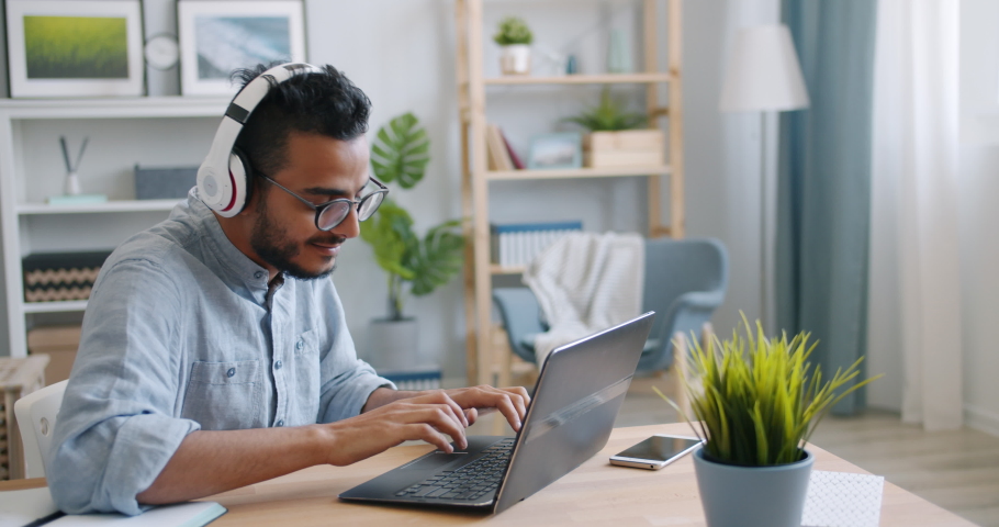 Slow motion of African American man using laptop and listening to music in headphones at home relaxing and working at table. People and leisure concept. | Shutterstock HD Video #1036319159
