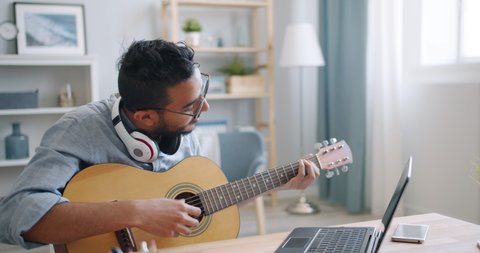 Young Arab guitarist is playing the guitar and using laptop typing at home alone enjoying leisure time in apartment. Hobby, music and youth concept.