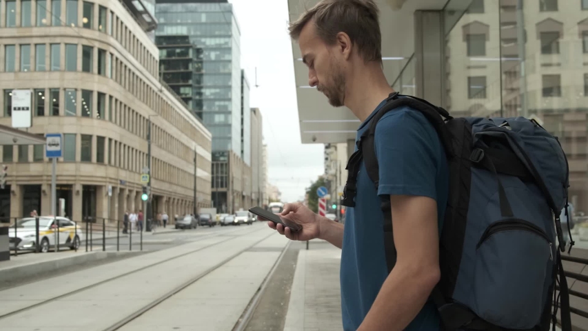 Millennial man tourist with backpack and smartphone stand on public transport stop and waiting tram in modern city center. Travel concept. Royalty-Free Stock Footage #1036327883