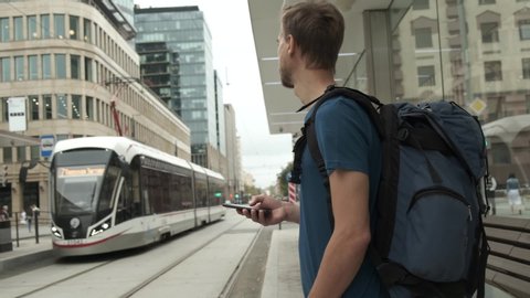 Millennial man tourist with backpack and smartphone stand on public transport stop and waiting tram in modern city center. Travel concept.