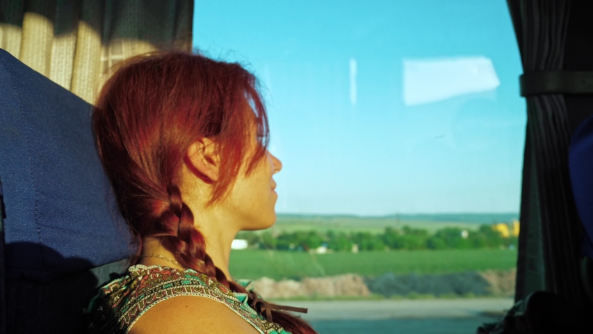 A lonely red-haired traveler girl looks out the bus window at road transport, nature and the blue sky. Summer, sunset time.