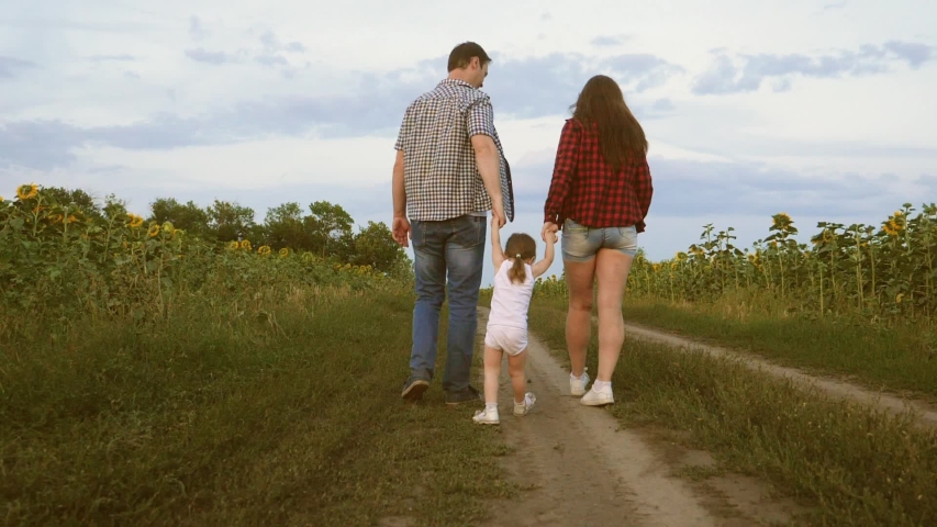 child mom and dad play together outside city in nature. little daughter jumping holding hands mom and dad. Family with small child walks along road and laughs next to field of sunflowers.