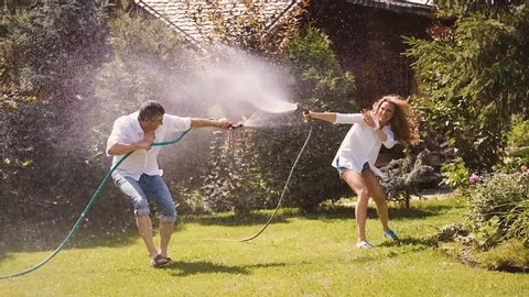 A married couple of young people in the backyard of a country house plays watering each other from a hose.