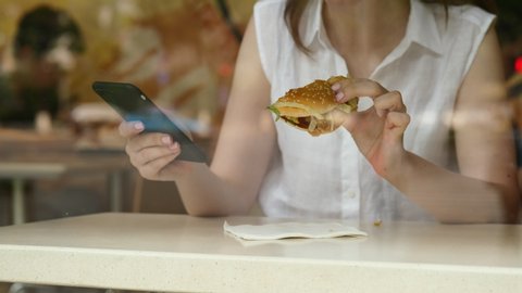 Young woman eat hamburger and browse smartphone, sitting in fast food restaurant face to window. Counter table and hands through glass, some street reflection. Business district area at Chinese city