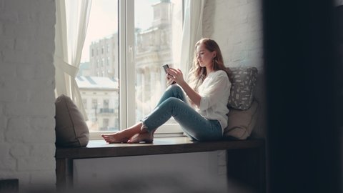 Charming young woman sitting on a windowsill at home and texting on her phone communication female looking message cellphone cheerful smile use internet modern smartphone portrait slow motion