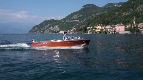 Como Lake September 3, 2019: Cinematic view of a person driving a luxury motor boat made of wood on Lake Como against the background of buildings near the water. Romantic sunset journey. Riva boat