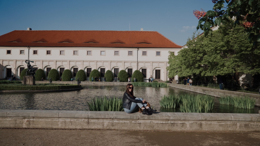 Pretty young lady with backpack sitting on parapet of pond. There are Wallenstein Palace and the pond with fish and sculptures on background, currently the home of the Czech Senate in Prague, Czech