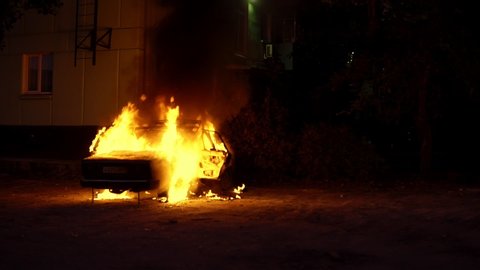 Exploding burning car on fire in front of building. Theme of riots criminal arson of a car near house. Automobile engulfed in flames of fire. Trees close to burning sedan.