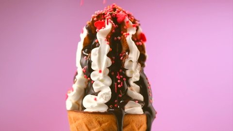 Ice cream cone close-up. Colorful sprinkles falling on ice cream with chocolate syrup topping. Icecream in waffle cone, rotating on pink background. White Sweet dessert closeup, rotation. 4K UHD