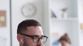 Tilt down of Caucasian man wearing glasses and wireless headphones sitting at laptop, communicating with client by video link while his colleagues working on background