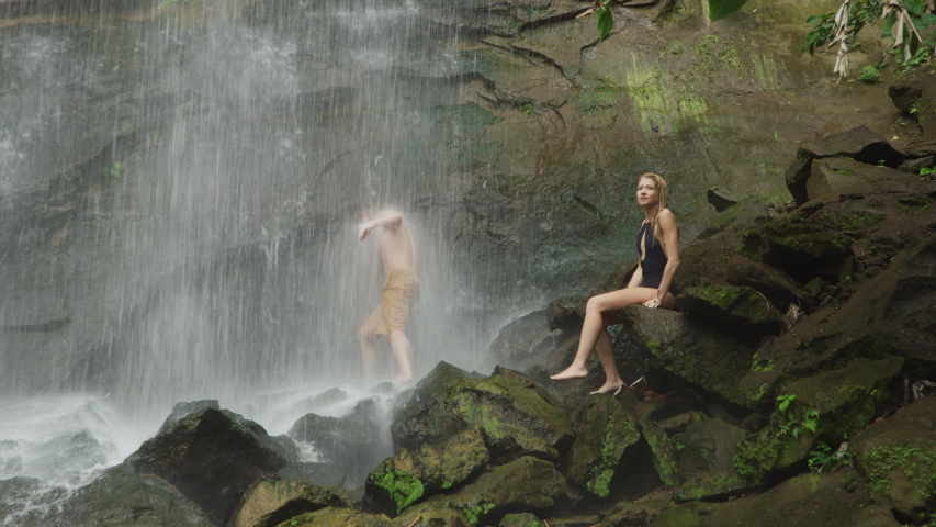 Woman sitting on rock watching water splashing on man standing under waterfall / Carmel Falls, Grenada