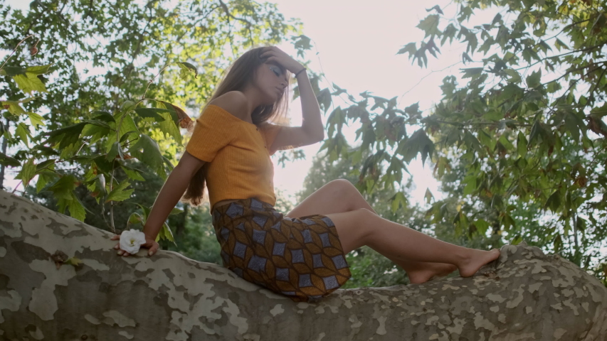 Tracking shot of attractive brown haired girl sitting on tree branch thoughtfully looking around in old garden at sunset