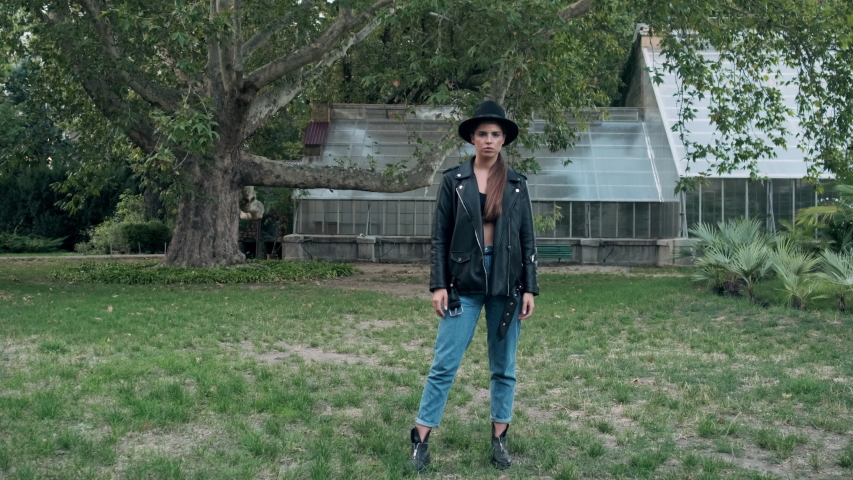 Tracking in shot of serious girl in hat and leather jacket crossing hands sadly looking in camera with greenhouse on background in old garden