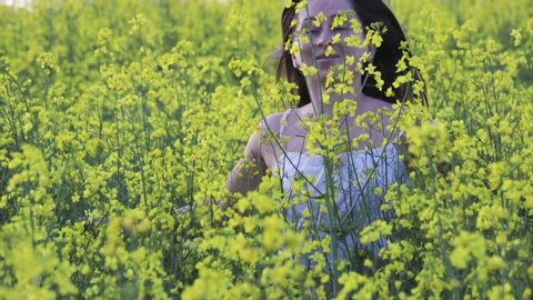 Sexy young girl in short white dress runs in a field with yellow rape flowers, her dark hair flying in the wind, slow motion