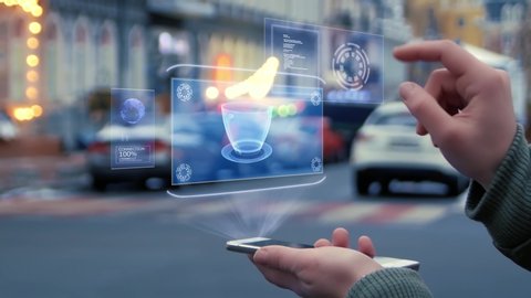 Female hands on the street interact with a HUD hologram with cup of coffee. Woman uses the holographic technology of the future in the smartphone screen on the background of the evening city