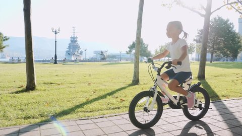 A little blonde girl is riding a bike along the seafront against the sun. A small child is moving at the camera and action changing to the side view of the bike and a kid.