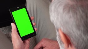 An elderly man sits on a couch in an apartment and looks at a smartphone with a green screen - closeup from behind his head