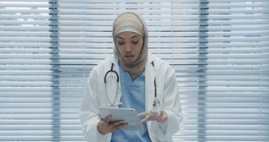 Animation of a portrait of a young mixed race female doctor wearing a hijab looking to camera holding a tablet computer and talking, seen on a screen of a digital camera in record mode with icons and