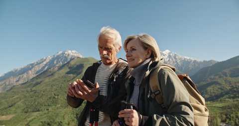Senior caucasian couple having a nordic walk in mountains, then stopping to take a picture with smartphone. Old people traveling together after retirement - pension, tourism concept 4k
