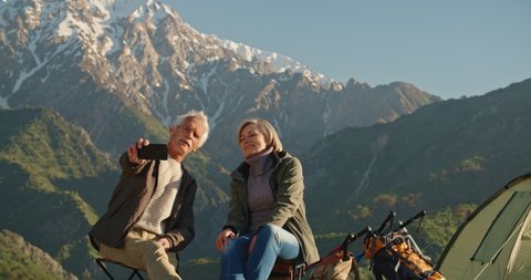 Senior cacasian couple having a rest on top of a mountain, taking a picture or having video chat on smartphone, traveling together after retirement - pension, recreational pursuit, tourism concept 4k