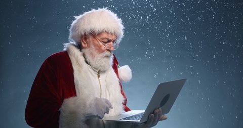 Santa Claus holding laptop in hands, typing, then looking at camera and winking, isolated over blue background - christmas spirit concept close up 4k footage
