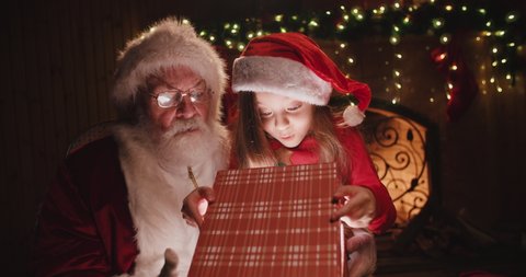 Santa claus sitting on his rocker with little caucasian girl sitted on his knee, opening up a gift with something special together - christmas spirit, holidays and celebrations concept 4k footage