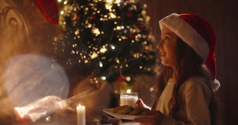 Little caucasian girl wearing christmas hat leaving cookies and milk near chimney for santa - holidays and celebrations, christmas spirit concept close up 4k