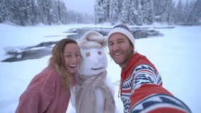 Young couple having fun outdoors in the snow taking selfies with snowman surrounded baby winter wonderland. Couple taking cool selfies in winter 