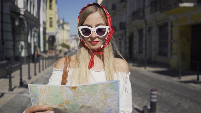 Positive beautiful young woman standing on the street with the map in hands. Attractive fashionable girl trying to find a way in the old European city. Tourism concept, vacation