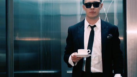 Attractive stylish businessman wearing sunglasses comes out of the elevator with a cup of coffee in his hands. A man in a jacket and tie goes to his office.