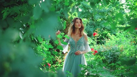 A woman in a luxurious evening dress of turquoise color walks in the garden among blooming roses. Princess with stylish styling on her loose hair. Fashionable image of a graduate girl at a prom party