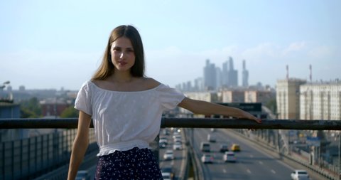 young pretty townswoman is posing for camera on a bridge over road and cityscape in distance
