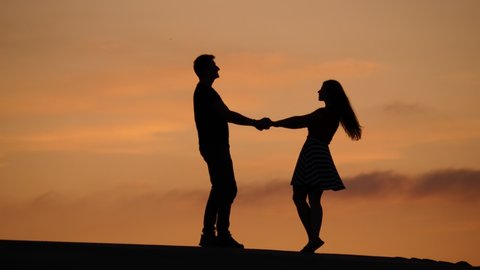 Lovely couple holding hands and turn around, two people dance at sunset. Silhouetted shot against neat yellow sky, nice summer evening time