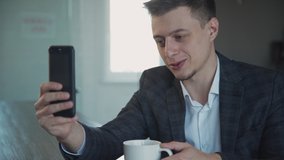 Young american businessman having video call holding coffee in hand in office desk. Portrait of happy man watching smartphone screen and talking with smile during working day in company. Concept