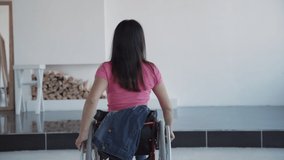 Adult disabled female wearing in denim jeans and casual clothes sitting in special invalid chair or handicap inside bright house with light room. Young woman in wheelchair spending weekend time home