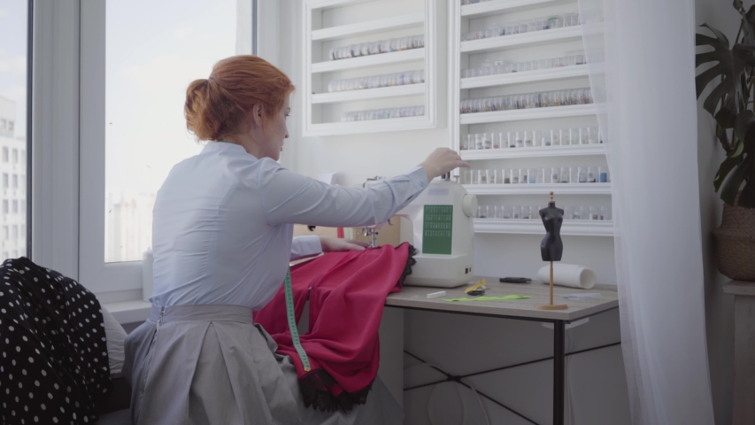 Successful cute red-haired woman sews at the table in workshop. Seamstress works diligently and carefully. Hobby and work. Sewing clothes. Back view. | Shutterstock HD Video #1036514690