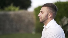 Close-up view of handsome bridegroom with brown hair and beard in white shirt looking at the camera and smiling in park. Action. Wedding video