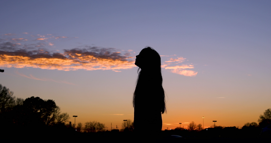 Stunning silhouette footage of young girl with long hair standing in a parking lot during a colorful sunset as she blows outwardly and chuckles to herself.